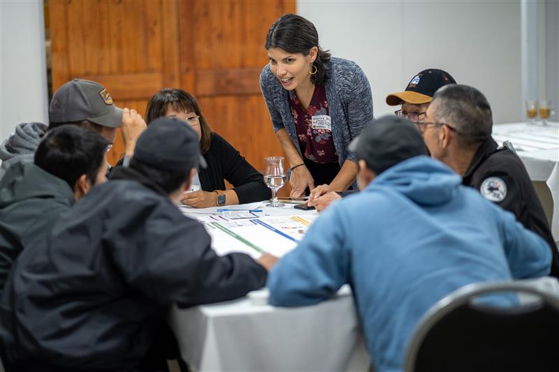 A standing woman speaks to people seated at a circular table as they lean forward to listen.