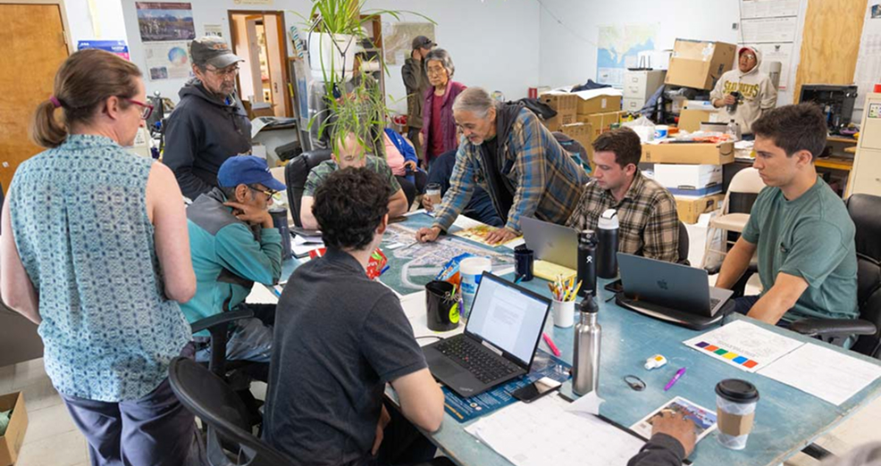 Nine people gather around a table with many papers as one older man points at a map and speaks to the group.